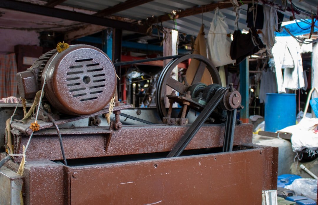 A close-up photograph captures an aged, heavy-duty industrial electric motor with a connected pulley and belt system, likely powering a piece of laundry machinery such as a washing machine or centrifuge.
